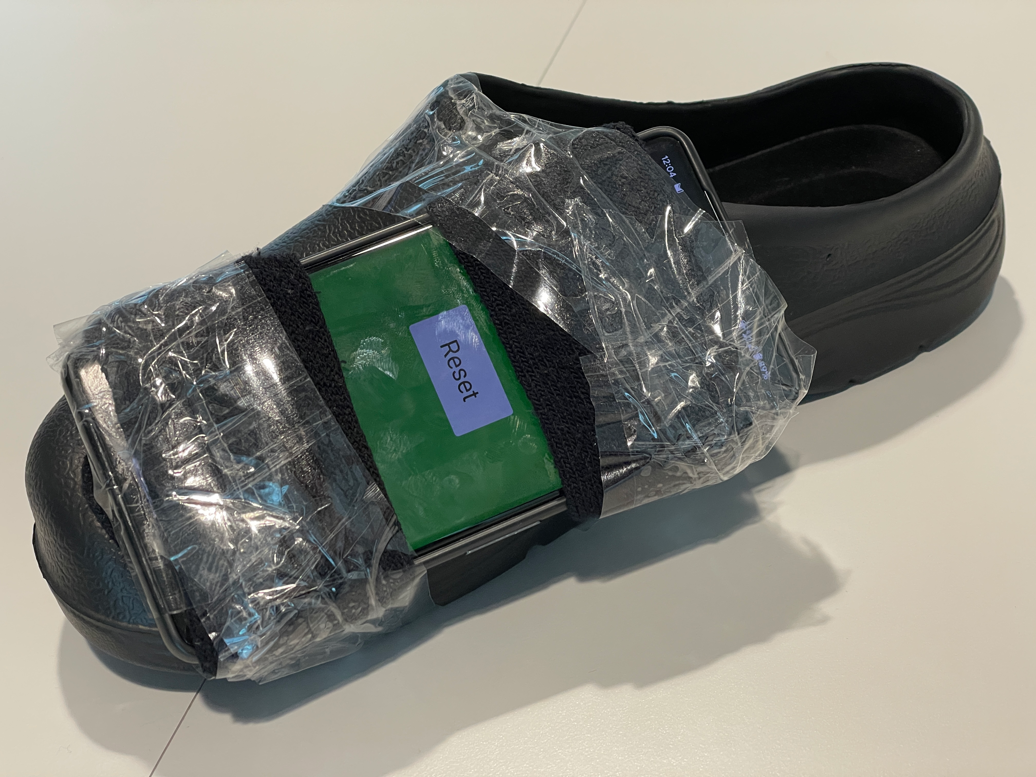 A single black foam slipper, with a smartphone taped to the inward facing side of the sole. The screen of the smartphone is facing up, displaying a simple interface with a button labeled 'Reset' and a green background.